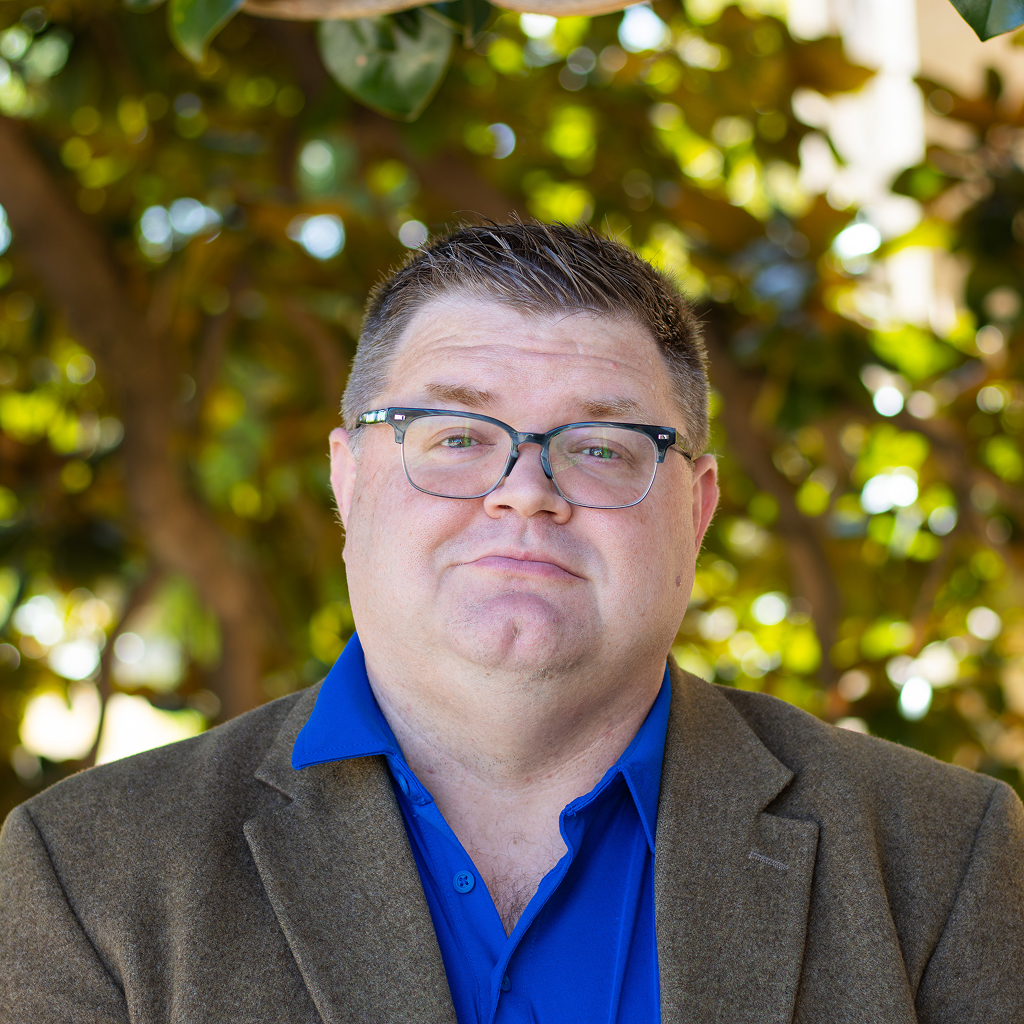 A man with glasses and blue button up shirt under brown suit 