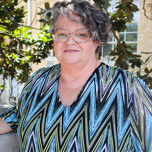 Woman in light blue, black, and white herringbone patterned shirt standing in front of a tree on campus