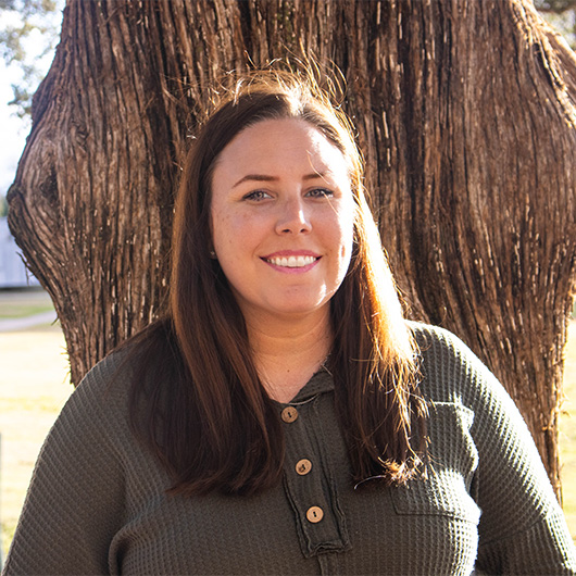 Woman with brown hair in a grey shirt, standing in front of a tree