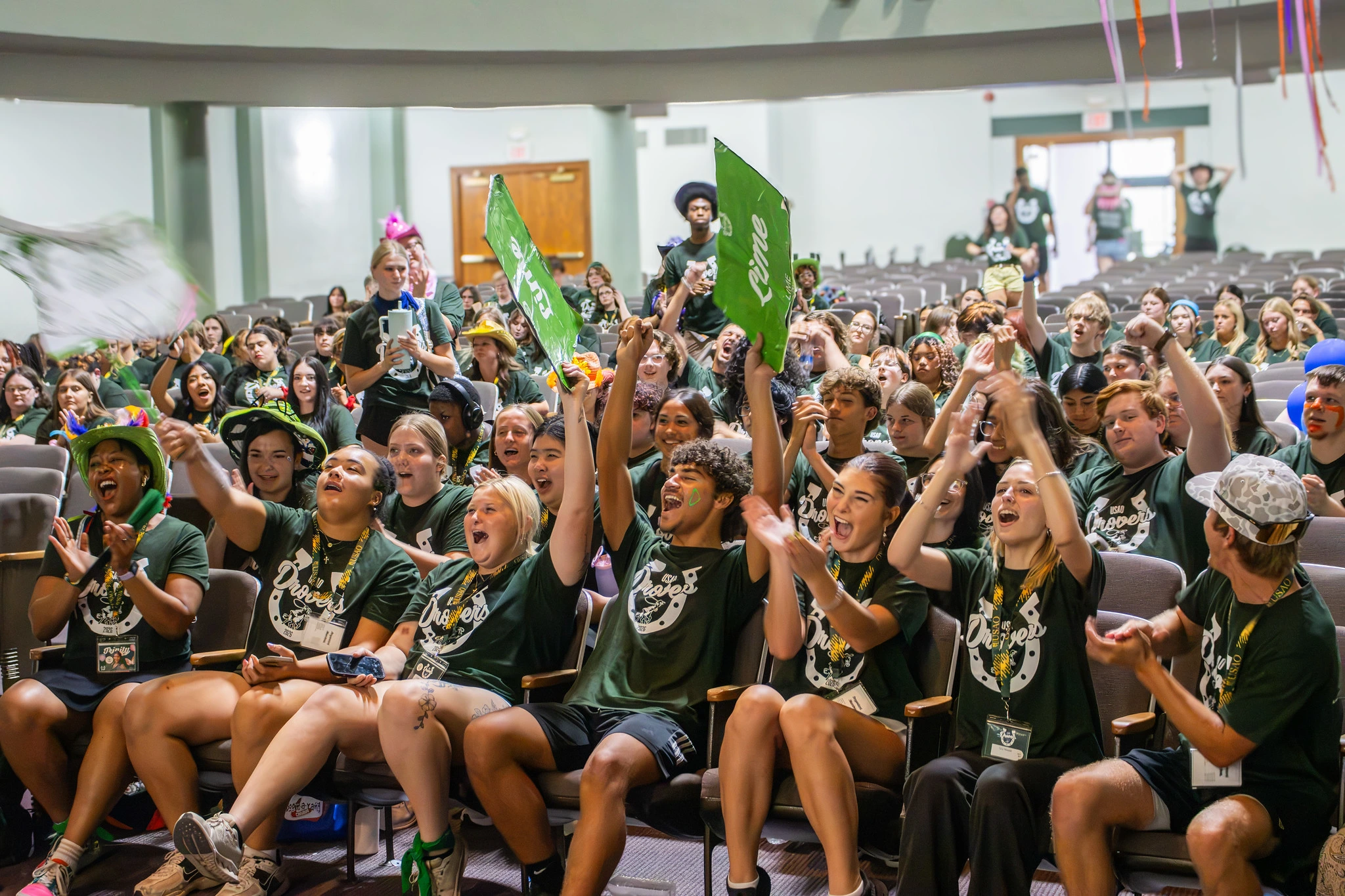 USAO students cheer, clap and wave green flags during a lively campus event in an auditorium.
