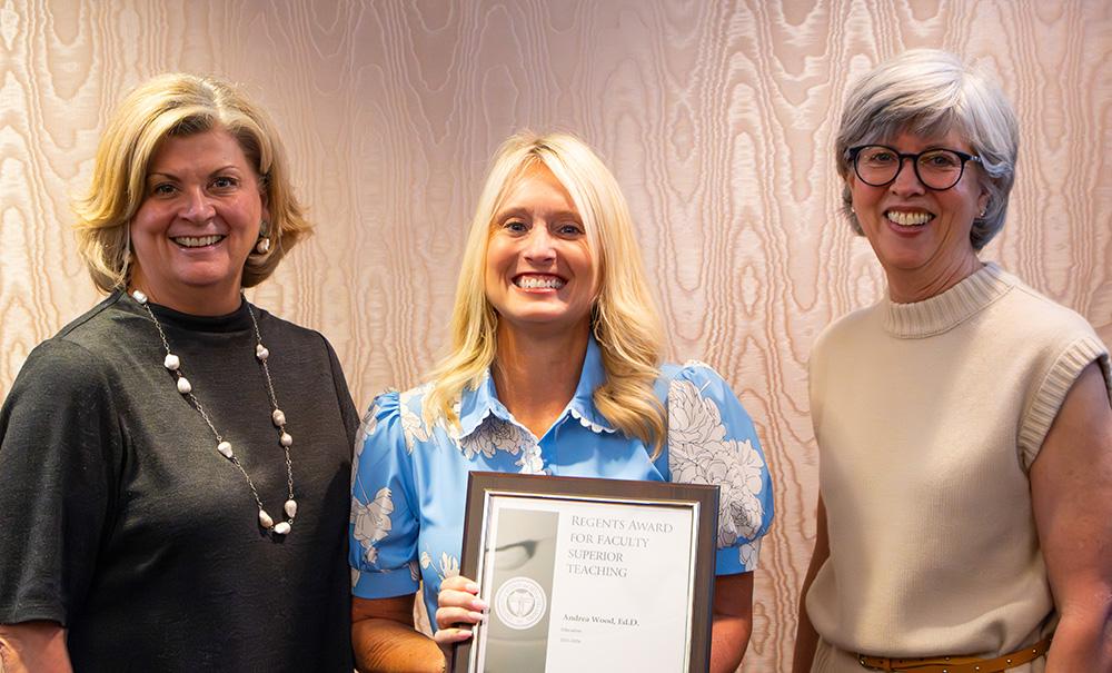 three people stand smiling with one in the middle holding an award